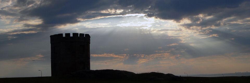A round stone tower is silhouetted against a dramatic sky, as sun rays—symbolic of hope after trauma—shine through breaks...