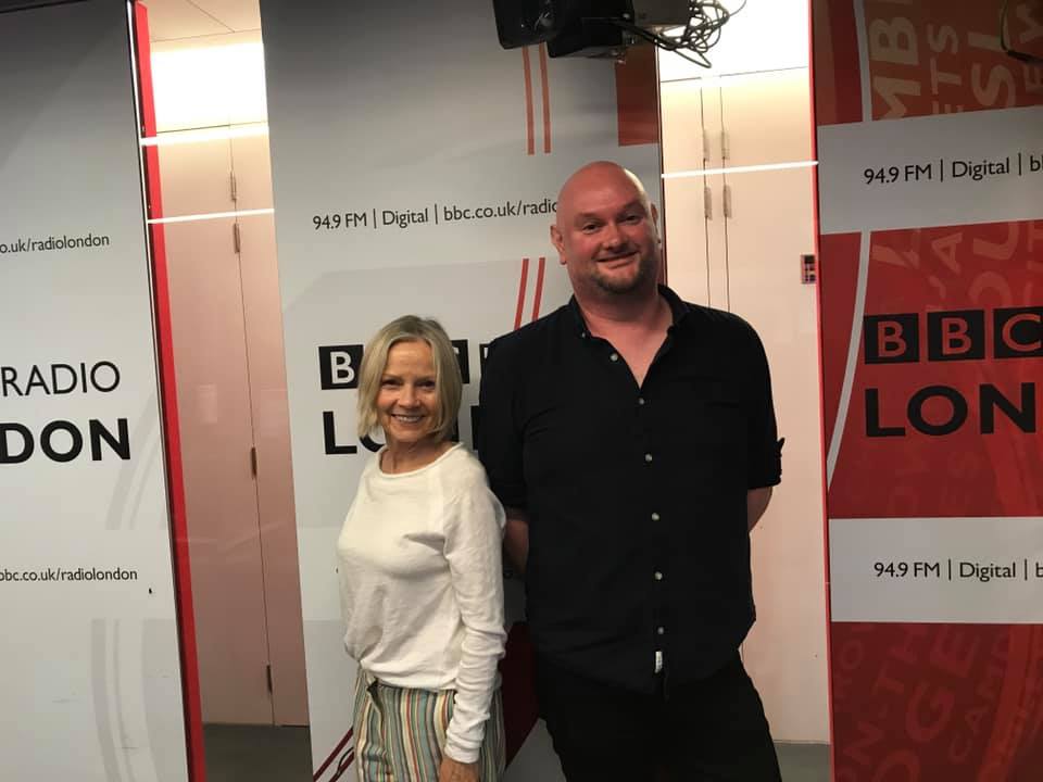 Two people stand smiling in a radio studio with BBC Radio London banners behind them. The woman, in a white top and...