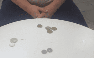 A person sits at a white round table with several coins scattered on its surface, hands clasped together in their lap—a...