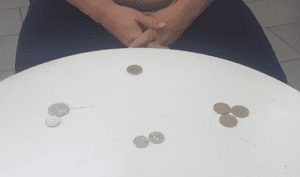A person with hands clasped sits at a white circular table, quietly grouping coins—a subtle reflection of trauma or PTSD....