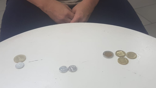 A person sits at a white round table with several coins spread out in three clusters, their hands resting in their lap—a...