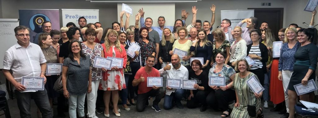 A large group of adults, smiling and holding certificates, pose together in a classroom or office setting. Some people...