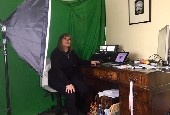 A woman sits at a wooden desk in front of a green screen with studio lighting. She faces a computer and laptop, preparing...
