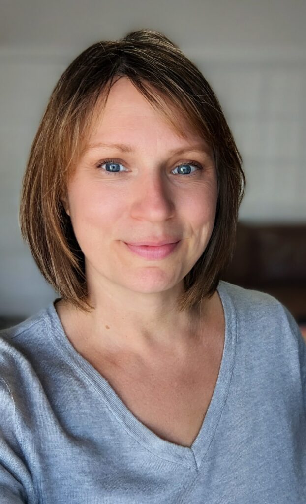 A woman with short, light brown hair and blue eyes is smiling softly. She is wearing a light grey V neck top and stands...