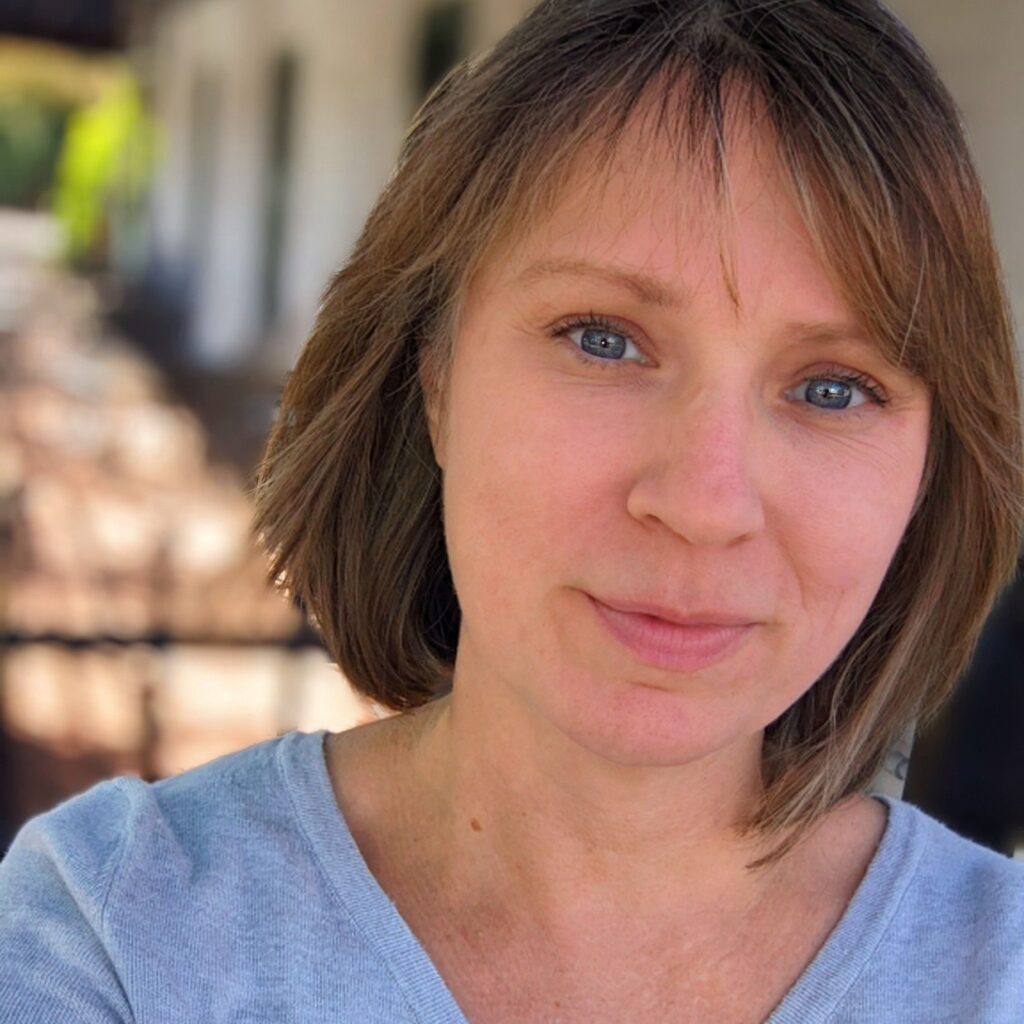 A woman with light brown hair and blue eyes, wearing a light blue top, stands outdoors with a blurred background of a building and trees. She looks into the camera with a slight smile, reflecting resilience after experiencing trauma.